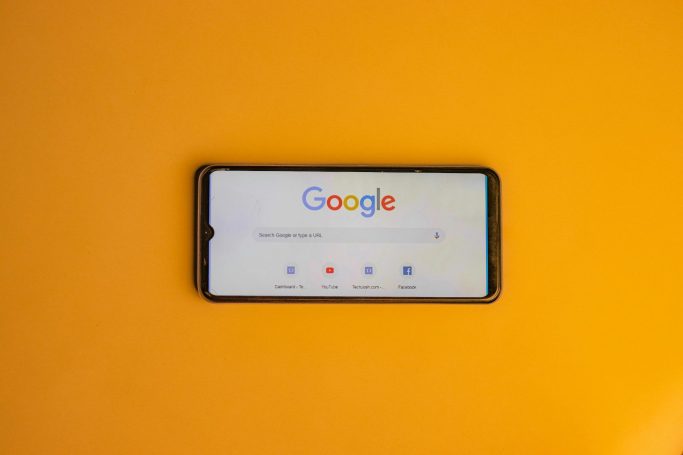 Smartphone mit Google-Suchseite vor orangefarbenem Hintergrund.